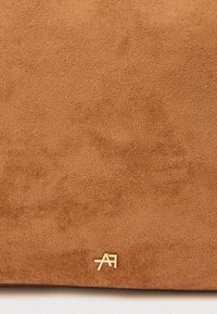 Τσάντα ώμου από ταμπά σουέτ με απαλή υφή, που διαθέτει χρυσό λογότυπο στη γωνία στο κάτω μέρος. Η επιφάνεια έχει μια λεία, διακριτική τελείωμα.