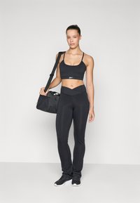 Woman in black sports bra and leggings stands holding a black gym bag over her shoulder against a plain light background.