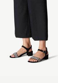 Sandalias negras con una correa de tela con estampado, hebilla en el tobillo y un tacón bajo de bloque. El diseño presenta acentos geométricos en blanco y negro.