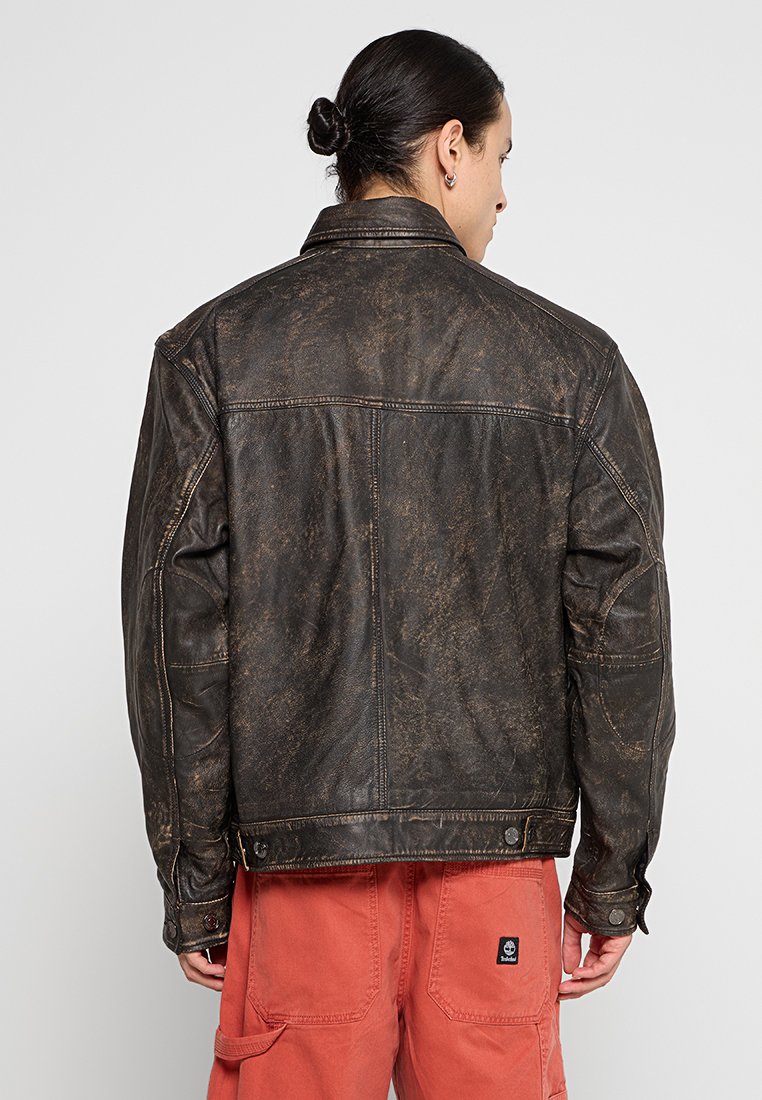 Brązowa, zniszczona skórzana kurtka o klasycznym kroju, widocznych szwach i zapinanych mankietach. Matowa tekstura i subtelne różnice w kolorze w całej kurtce.