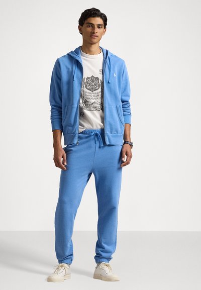 Hanorac bleu deschis și pantaloni jogger asortați, amândouă dintr-un material moale. Hanoracul are fermoar și buzunare frontale, iar sub el se află un tricou grafic alb.