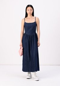 Denim maksi haljina sa tankim trakama, naboranim strukom i raširenom suknjom. Nosila s bijelim tenisicama i drži rozu clutch torbicu.