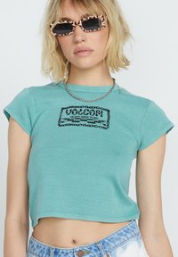 Turkoois cropped t-shirt, korte mouwen, met een zwart grafisch ontwerp waarop "Volcom" staat met een kettingrand. Draag het met een zilveren kettinghalsketting en gestippelde zonnebril.