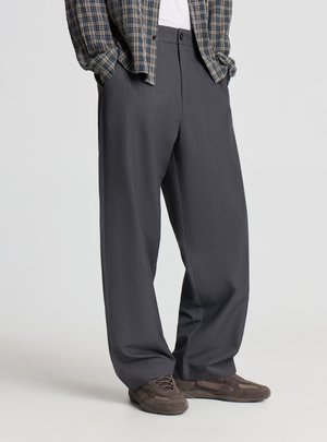 Uomo che indossa pantaloni larghi grigio scuro, sneakers marroni e una camicia a quadri con le mani nelle tasche, in piedi su un pavimento grigio chiaro.