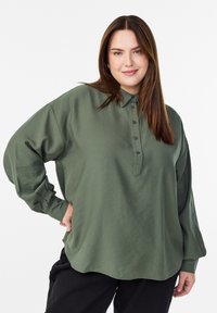 Kobieta z prostymi brązowymi włosami, ubrana w luźną oliwkową koszulę z guzikami i czarne spodnie, uśmiechająca się z jedną ręką na biodrze.