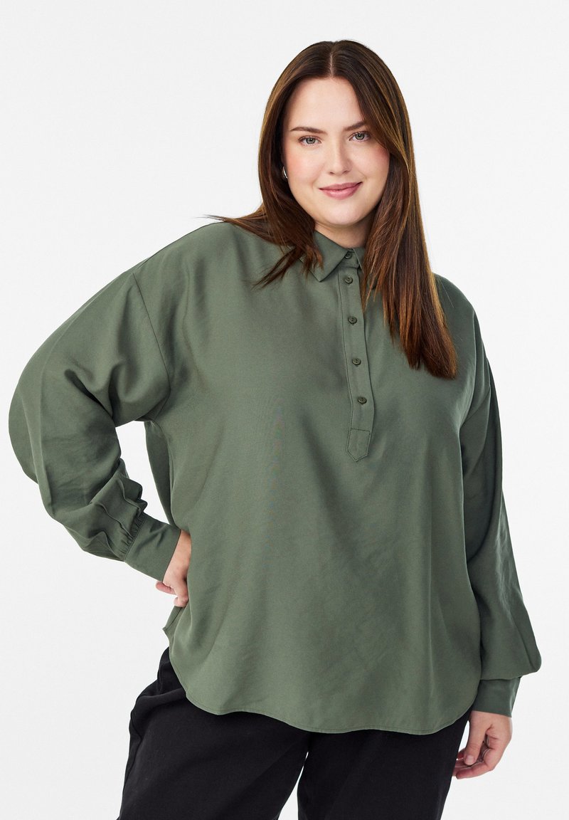 Kobieta z prostymi brązowymi włosami, ubrana w luźną oliwkową koszulę z guzikami i czarne spodnie, uśmiechająca się z jedną ręką na biodrze.