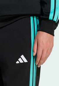 Pantalon de sport noir avec des bandes latérales turquoise, arborant un logo triangulaire blanc. Le tissu semble lisse ; les bandes ont une texture brillante.