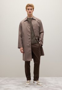 Cappotto lungo marrone chiaro con bottoni, maglione marrone testurizzato, pantaloni marroni e sneakers chunky bianche con dettagli beige. Sullo sfondo neutro.