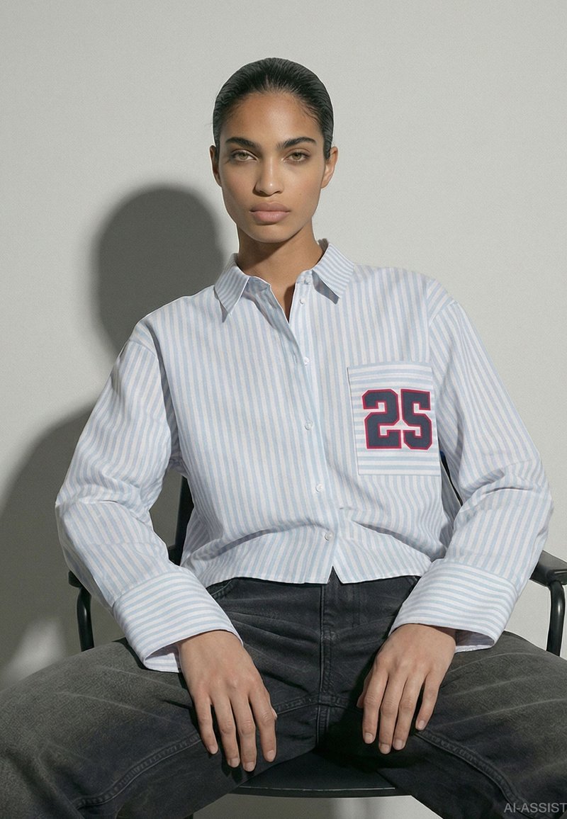 Femme aux cheveux foncés portant une chemise rayée avec le numéro 25 sur la poche, assise, jambes écartées et mains posées sur les cuisses sur un fond gris.