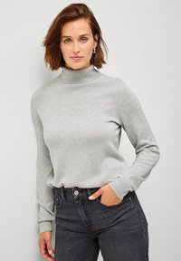 Maglione a collo alto grigio chiaro in tessuto morbido, con maniche lunghe e polsini a coste, abbinato a jeans scuri. Design semplice e classico.