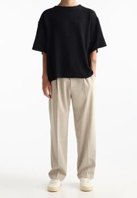 Personne portant un haut noir ample à manches courtes, un pantalon beige à jambes larges, des baskets blanches, et un tatouage visible sur l'avant-bras, sur fond blanc.