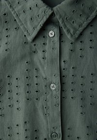 Chemise vert foncé avec un col, boutonnage à l'avant et motif de broderie à œillets avec de petites ouvertures sur l'ensemble du tissu.