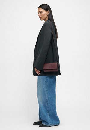 Woman wearing a dark oversized blazer, wide-leg blue jeans, black pointed shoes, and a burgundy crossbody bag standing in profile.