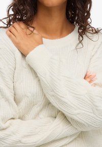 Γυναίκα με κρεμ πλεκτό πουλόβερ, με τα χέρια σταυρωμένα, το ένα χέρι να ακουμπά στον αντίθετο ώμο, με σγουρά σκούρα μαλλιά.