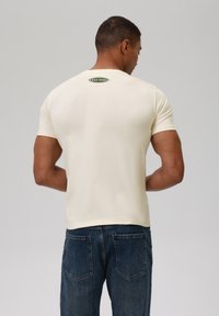 Camiseta corta de manga corta color crema con cuello redondo y una pequeña etiqueta verde que dice "EST 1958" en la parte superior de la espalda. Combinada con jeans de mezclilla azul.