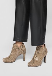 Bottes de cheville en simili cuir beige avec un motif serpent, dotées d'une fermeture éclair et d'un talon haut effilé, posées sur un pantalon noir.