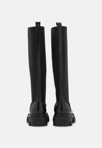 Botas negras hasta la rodilla con un tacón de cuero liso y elegante y un asta de tela texturizada. Presentan una suela de goma ligeramente elevada. Dos tiradores.
