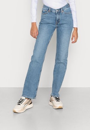 Partie inférieure du corps d'une personne portant un jean droit bleu clair, des baskets blanches avec des lacets noirs et un haut blanc à manches longues.
