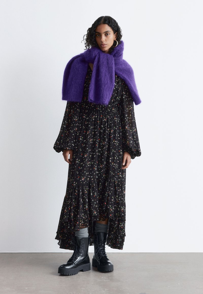 Langes schwarzes Kleid mit Blumenmuster, mit Rüschensaum, kombiniert mit einem lila, flauschigen Schal und schwarzen Plateau-Stiefeln. Glatte Stoffoberfläche mit strukturierten Akzenten.