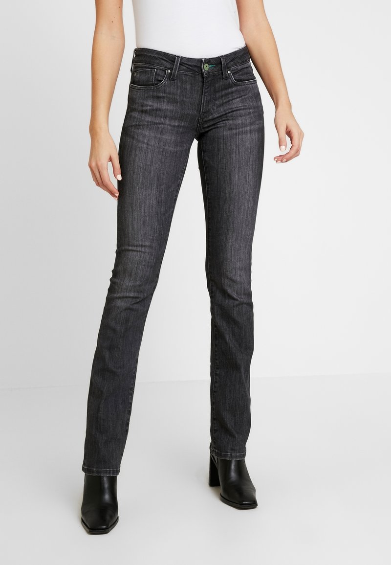 Femme portant un jean slim gris foncé et des bottines noires à talons sur un fond blanc uni.