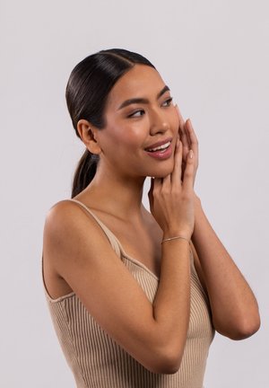 Tan ribbel tanktop met een brede halslijn, gedragen door een persoon met een zilveren armband, tegen een effen lichte achtergrond. Het haar is naar achteren gestyled.