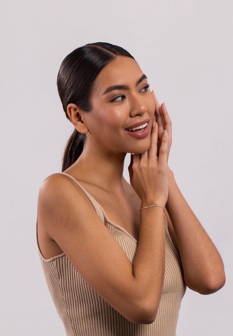 Beige gerippter Tanktop mit weitem Ausschnitt, getragen von einer Person mit einem silbernen Armband, vor einem schlichten hellen Hintergrund. Die Haare sind zurückfrisiert.