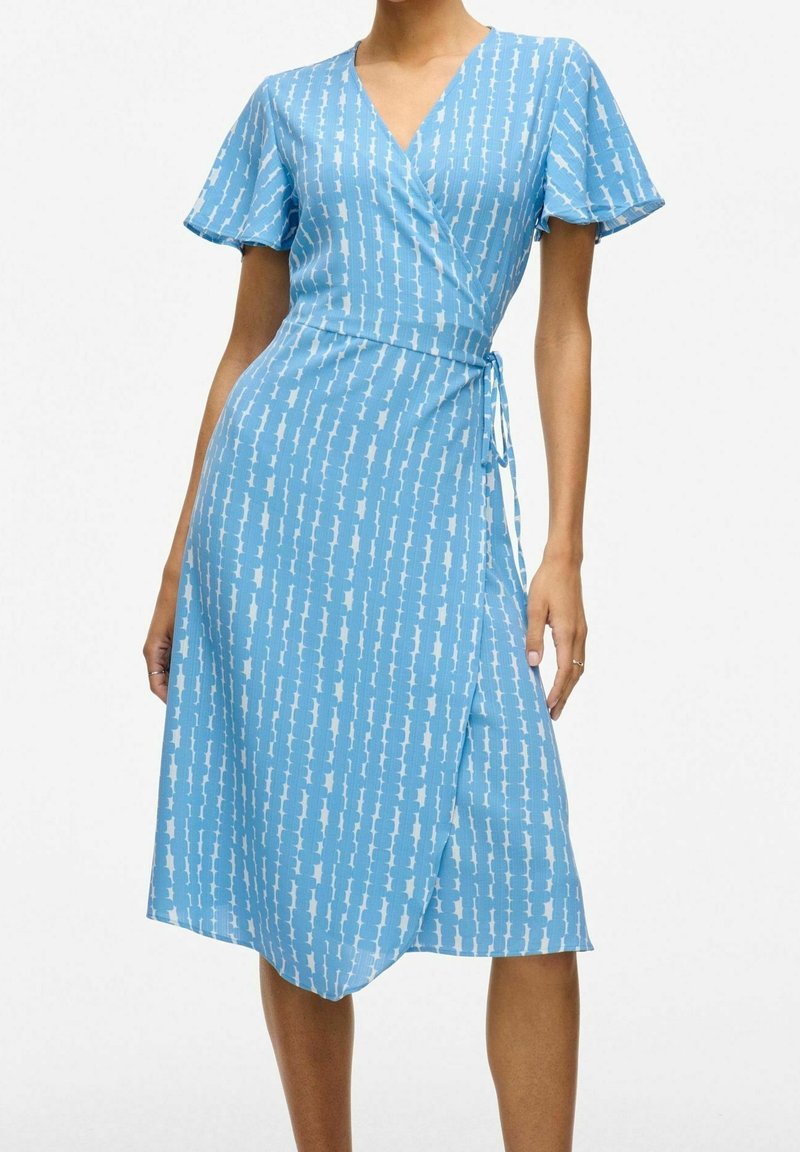 Femme portant une robe portefeuille bleu clair jusqu'aux genoux avec des manches courtes flottantes et un motif abstrait blanc vertical.