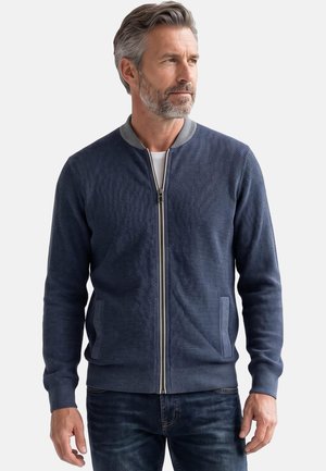 Mann mittleren Alters mit grauen Haaren und Bart, trägt eine strukturierte blaue Jacke mit Reißverschluss und dunkle Jeans, schaut zur Seite.
