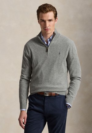 Homme portant un pull gris à fermeture éclair sur une chemise rayée bleue, un pantalon foncé et une ceinture marron, debout devant un fond uni.