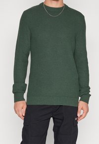 Pull en maille verte avec encolure ronde, finition texturée, poignets et ourlet côtelés. Associé à un pantalon cargo foncé pour un contraste.