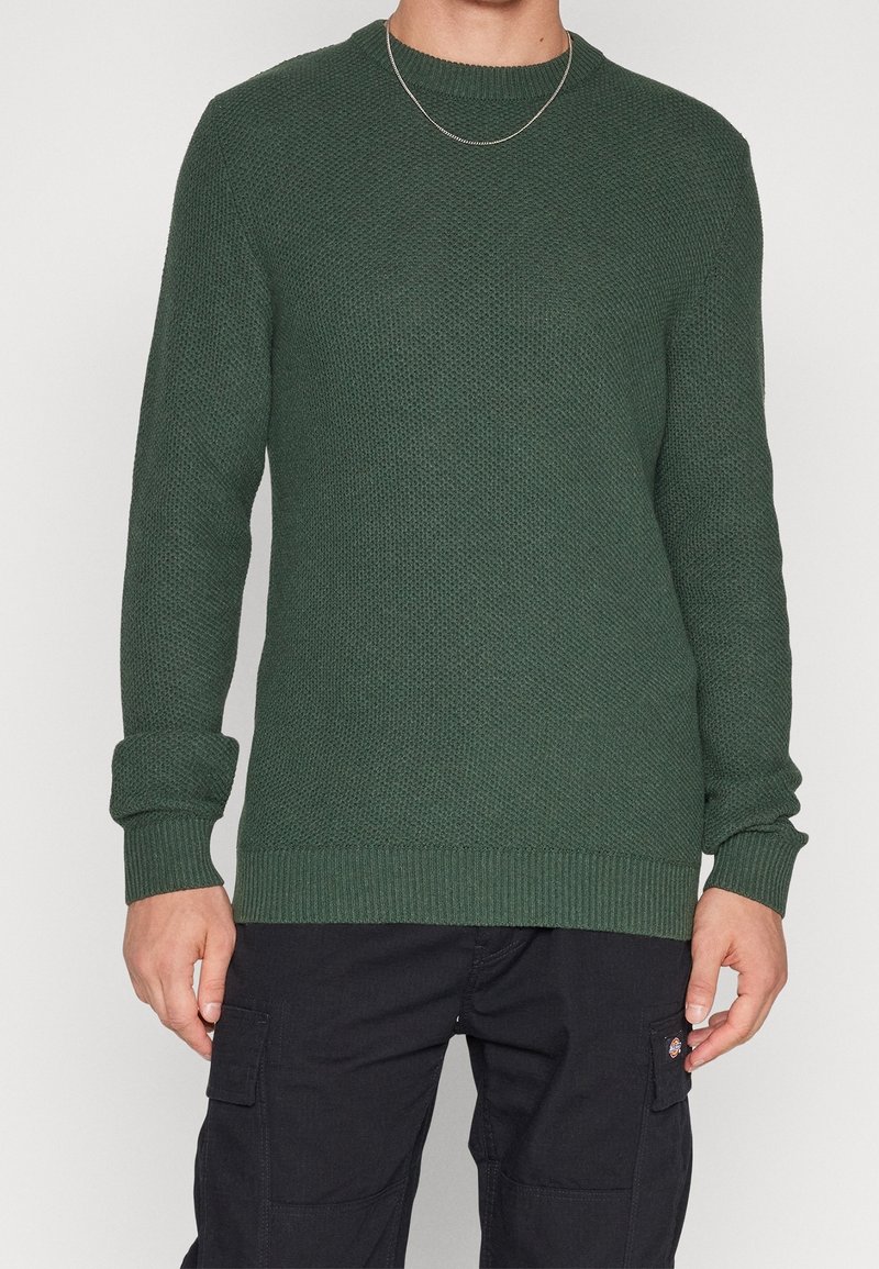 Pull en maille verte avec encolure ronde, finition texturée, poignets et ourlet côtelés. Associé à un pantalon cargo foncé pour un contraste.