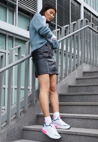 Mladá žena v modrém svetru, černé kožené sukni a bílých teniskách s růžovými akcenty stojí na městských kamenných schodech.