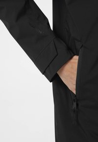 Černá voděodolná bunda s kapsou na zip a označeným rukávem. Materiál je hladký a matný, s přizpůsobeným střihem pro funkčnost.