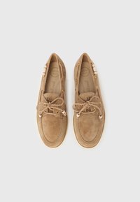 Par de mocassins de camurça bege com laços de fita e detalhes de contas, rotulados como "super macios" no interior, sobre um fundo branco.