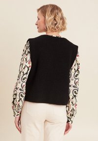 Schwarzes, gestricktes, ärmelloses Oberteil mit einer strukturierten Oberfläche, darunter eine gemusterte Bluse mit langen Ärmeln in Creme-, Grün- und Rottönen.