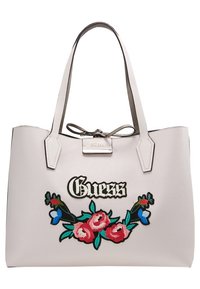 Sac fourre-tout rose clair avec deux poignées, fleurs rouges et bleues brodées, et logo "Guess" en lettres noires et blanches sur le devant.