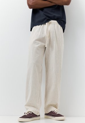 Man wearing beige pinstripe wide-leg pants with drawstring waist, black t-shirt, and dark brown sneakers with white soles.