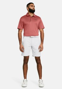 Rotes Polo-Shirt mit Kragen, kombiniert mit weißen Shorts. Mit einem Logo auf der linken Brust und einem Gürtel. Trägt weiße Golfschuhe.