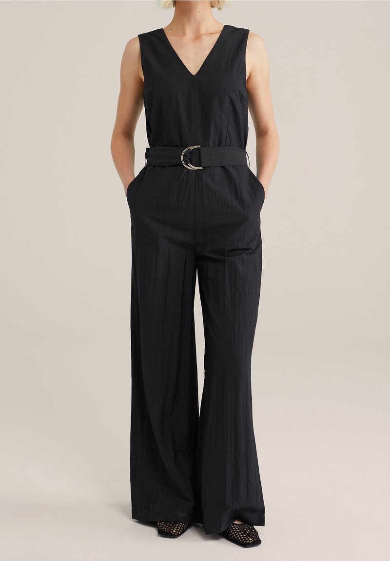 Femme portant une combinaison noire sans manches avec un col en V, des jambes larges, des poches latérales et une ceinture en tissu avec une boucle double en métal.
