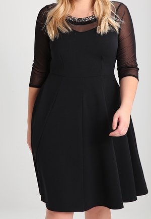 Kobieta w czarnej sukience do kolan z przezroczystymi rękawami do łokcia oraz zdobieniem w postaci perełek przy dekolcie.