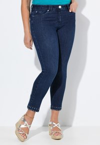 Tmavě modré džíny skinny mají střední pas, přiléhavý siluetu a kovové akcenty na okraji kotníku. Jsou kombinovány s kovovými platformovými sandály.