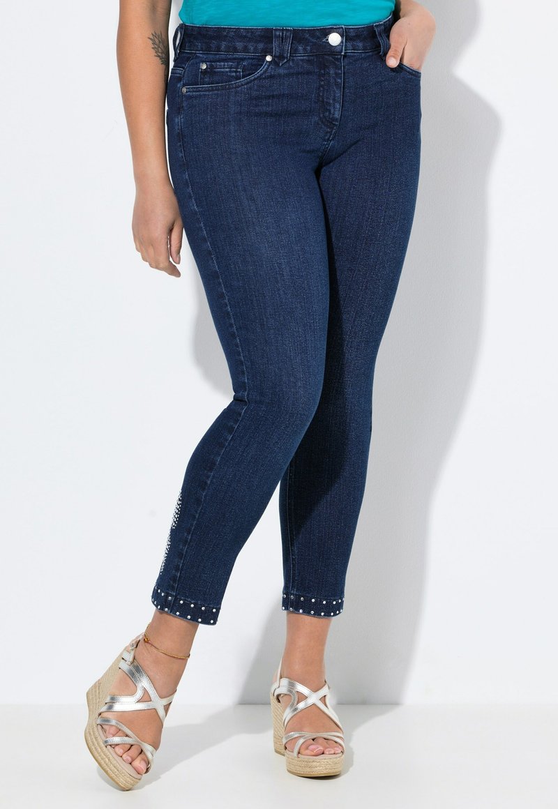 Tmavě modré džíny skinny mají střední pas, přiléhavý siluetu a kovové akcenty na okraji kotníku. Jsou kombinovány s kovovými platformovými sandály.