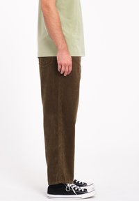 Pantalon en velours côtelé marron avec des côtes verticales, coupe droite, poches latérales, porté avec un t-shirt vert clair et des baskets noires.
