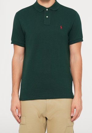 Man die een donkergroen poloshirt draagt met een rood geborduurd logo op de linkerborst en beige cargobroek, tegen een effen achtergrond.