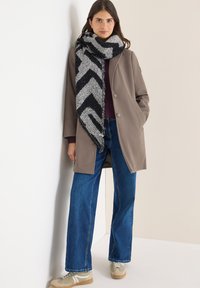 Brun ytterrock med stora knappar, bärs över en mörk tröja. Kombineras med blåvida byxor och en svartvit mönstrad scarf.