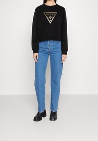 Sudadera negra con un logo triangular dorado, combinada con jeans azules de pierna recta y botas negras con tacón grueso. Diseño simple, ajuste casual.