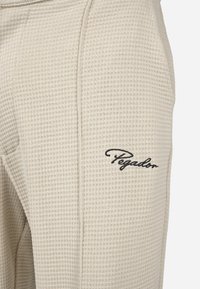 Beige bombažne hlače s skrčenjem, ki značilno prikazujejo teksturiran vzorec vaflčne pletenine, z izvezenim logotipom "Pegador" v črni barvi na strani.