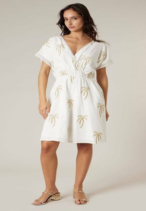 Femme portant une robe blanche à manches courtes avec des imprimés de palmiers dorés et des sandales à talons blocs dorés, debout devant un fond uni.