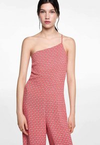 Punainen kuvioitu haalari, jossa on yksi olkaimi ja joka esittelee geometrisia muotoja sekä rentoa istuvuutta. Liukas kangas ja joustava vyötärö.