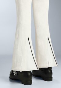 Fehér bordázott, bővülő nadrág oldalsó cipzárokkal és texturált anyaggal, fekete cipőkkel párosítva, melyeken fém díszítés és texturált talpak találhatók.